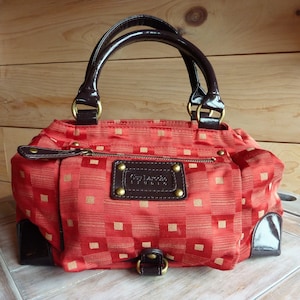 以下が含まれることがあります： 赤と金のチェック柄のハンドバッグ。ブラウンの革製のハンドルと、ブラウンの革のパッチに金色の文字で「Guy Laroche Studio」と書いてあります。