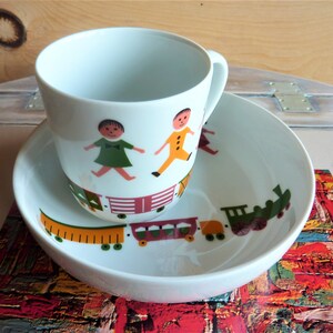 Puede incluir: Juego de taza y platillo de cerámica blanca para niños con un diseño de tren y niños. La taza tiene un asa y el platillo tiene un borde.