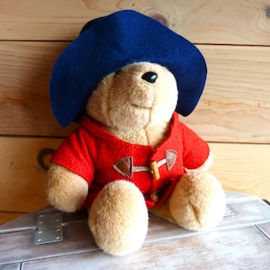 May include: A plush brown bear wearing a red coat and a blue hat. The bear is sitting on a wooden surface.