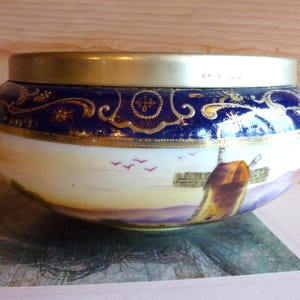 May include: A large, white porcelain bowl with a blue and gold rim and a painted scene of a windmill and birds. The bowl has a silver lid.