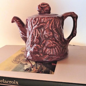May include: A glossy, dark brown ceramic teapot featuring a relief design of rabbits on the body and a rabbit-shaped finial on the lid. The teapot has a curved handle and a spout, with a textured surface that mimics the appearance of tree bark.