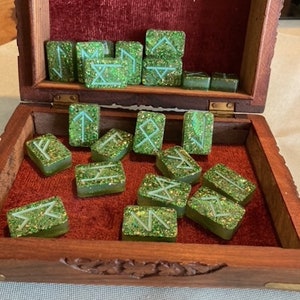 Può includere: Una scatola di legno con un interno in velluto rosso che contiene pietre runiche verdi con glitter. Le rune sono incise con linee bianche.