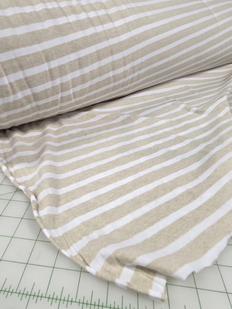 May include: Tan and white striped fabric, rolled and spread out. The fabric has a repeating pattern of horizontal stripes. The material appears to be a soft, woven textile, suitable for various sewing projects.