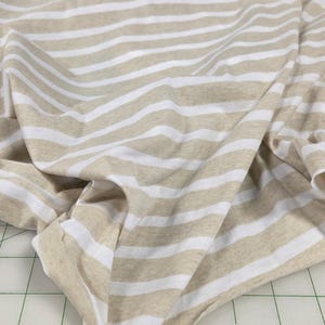 May include: Tan and white striped fabric. The fabric is folded and draped, showing the pattern of horizontal stripes. The stripes are evenly spaced and run across the material. The fabric appears to be soft and lightweight.