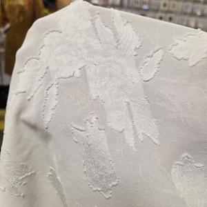 May include: Close-up of a white fabric with a raised floral pattern. The fabric appears to be semi-sheer, with the floral design creating a textured effect. The background is blurred, suggesting a retail environment.