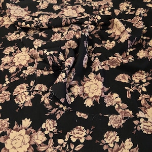 May include: A piece of black fabric featuring a repeating floral pattern of light beige roses and leaves. The material is draped to show folds and texture, displaying a consistent botanical print across the entire surface of the textile.