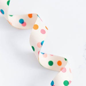 May include: A cream-colored ribbon with a repeating pattern of colorful polka dots in shades of orange, pink, green, and blue. The ribbon is curled and appears to be made of fabric. The background is white.