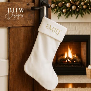 Può includere: Una calza di Natale bianca con il nome "EMILY" in lettere dorate è appesa a una porta marrone scuro. La porta ha una maniglia nera ed è leggermente socchiusa, rivelando un camino con fuoco acceso, candele e decorazioni festive.