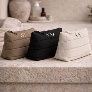 May include: Three quilted cosmetic bags in beige, black, and cream, each with a gold zip and monogram. The bags are arranged on a stone surface, with a neutral background featuring a vase and toiletries.