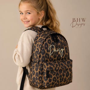 May include: A backpack featuring a brown and black leopard print pattern with the name Daisy written in gold script on the front pocket. The bag includes black zippers, black adjustable shoulder straps, and a front storage compartment.