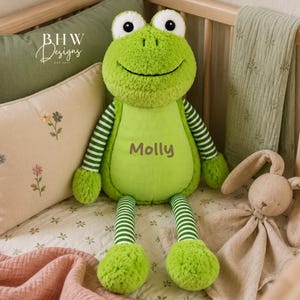 Könnte beinhalten: Ein Frosch-Stofftier mit leuchtend grünem Fell, weiß-grün gestreiften Armen und Beinen sowie großen weißen Augen mit schwarzen Pupillen. Auf der Brust ist der Name Molly in braunem Garn eingestickt. Das Spielzeug befindet sich in einem Kinderbett.