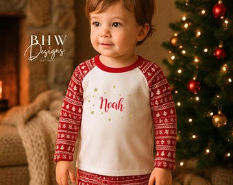 May include: A child wearing a red and white Christmas pajama set. The top is white with red sleeves and a red collar, featuring the name "Noah" and gold stars. The pants are red with a white pattern. A Christmas tree is in the background.
