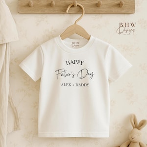May include: White t-shirt with the text "HAPPY Father's Day ALEX + DADDY" in black script. The shirt is hanging on a wooden hanger. A wooden rack with pegs is above the shirt. A stuffed bunny is in the lower right corner.