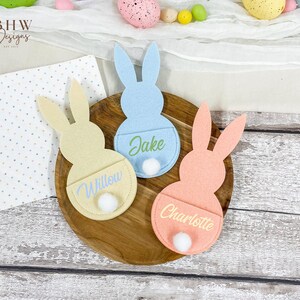 Op de afbeelding: Drie pastelkleurige vilten paasplaatskaarten in de vorm van een konijn met witte, pluizige staarten. Elk konijn heeft een zakje met een naam: "Willow", "Jake" en "Charlotte". De kaarten liggen op een houten dienblad, met paaseieren op de achtergrond.
