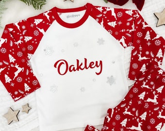 May include: A red and white Christmas pajama set with the name "Oakley" in red script. The long-sleeved top has red sleeves with a pattern of reindeer, snowflakes, and Christmas trees. The matching pants have the same pattern. The pajamas are a festive holiday item.
