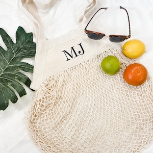 Puede incluir: Una bolsa de mano de malla de algodón blanco con una parte superior de lona de algodón natural. La bolsa tiene un monograma bordado en negro "MJ" en la parte delantera. La bolsa está llena de fruta, incluyendo una lima, una naranja y un limón.