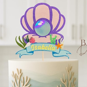 Op de afbeelding: Taart met twee lagen met een oceaanthema. De taart heeft een blauw-wit ombre-ontwerp, versierd met schelpen, koraal en zand. Een topper toont een parel in een schelp en de naam "Isabella" op een banner.