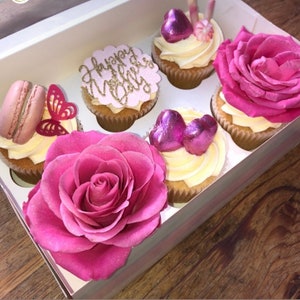 Puede incluir: Una caja de cupcakes decorados con rosas rosas, corazones rosas y un adorno de cupcake rosa que dice "Feliz D&iacute;a de la Madre".