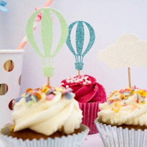 May include: Three cupcakes decorated with colorful sprinkles and topped with glitter hot air balloon and cloud cake toppers. The cupcakes are in white, pink, and brown cupcake liners.