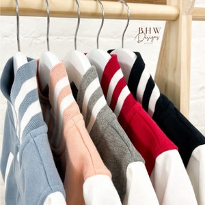 May include: Six different colored baseball jackets with white sleeves hanging on hangers. The jackets are blue, pink, gray, red, black, and white. The jackets have white stripes on the collars.