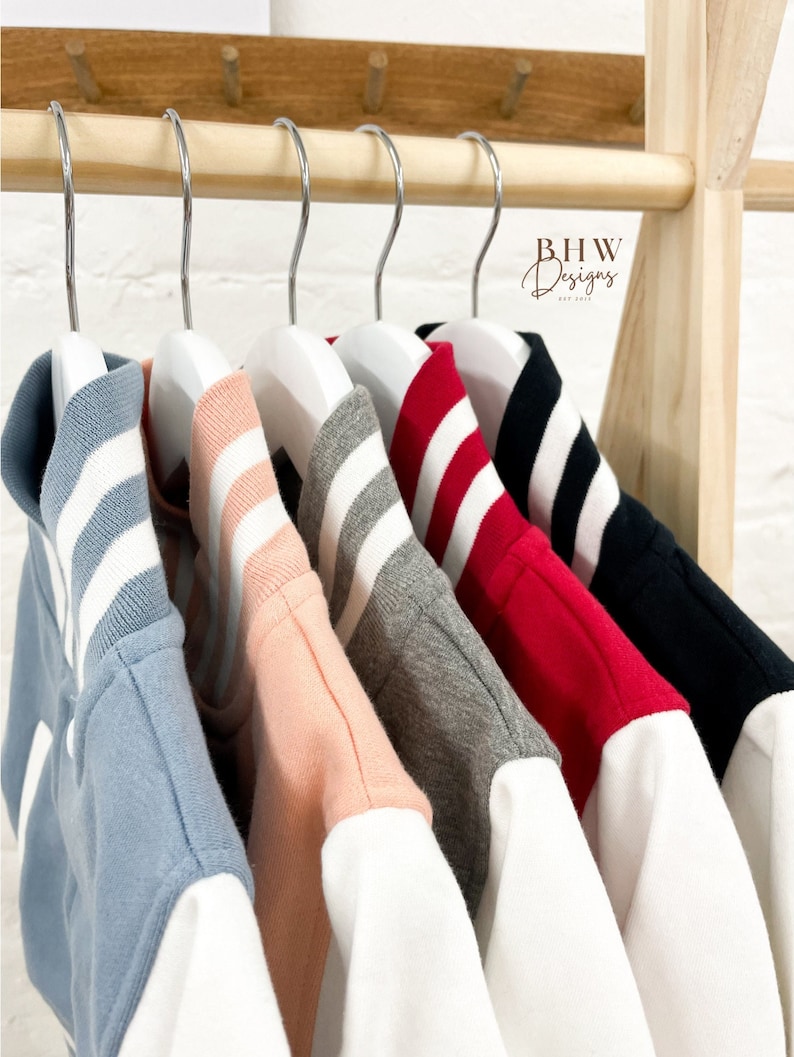 May include: Six different colored baseball jackets with white sleeves hanging on hangers. The jackets are blue, pink, gray, red, black, and white. The jackets have white stripes on the collars.