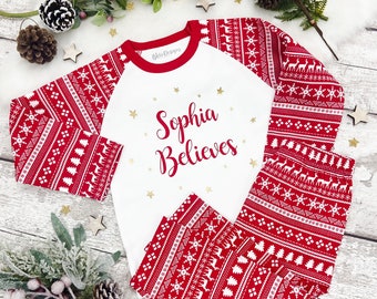 May include: A red and white Christmas pajama set with a festive pattern of reindeer, snowflakes, and trees. The white top features the text "Sophia Believes" in red script with gold stars. The pajamas are displayed on a wooden surface.