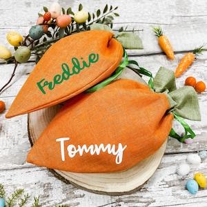 Puede incluir: Dos bolsas de tela naranja en forma de zanahoria con lazos verdes. Las bolsas están personalizadas con los nombres "Freddie" y "Tommy" en letras blancas.