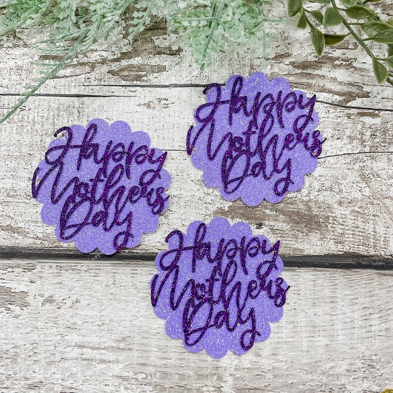 Puede incluir: Tres recortes de papel con purpurina de color morado con el texto "Happy Mother's Day" en una fuente cursiva.