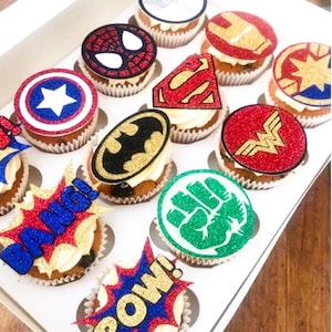 May include: A dozen cupcakes decorated with superhero logos and phrases like "POW!" and "BANG!" The logos are made of glitter and include Superman, Batman, Wonder Woman, Captain America, Iron Man, Spider-Man, Hulk, and Captain Marvel.