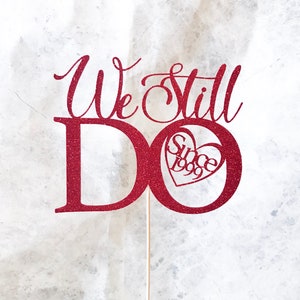 May include: Red glitter cake topper with the words "We Still Do" and a heart with "Since 1999" inside.