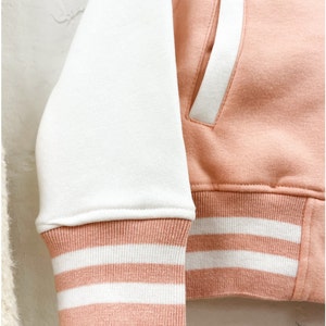 May include: A pink and white baseball jacket with white stripes on the cuffs and waistband. The jacket has a white pocket on the right side.