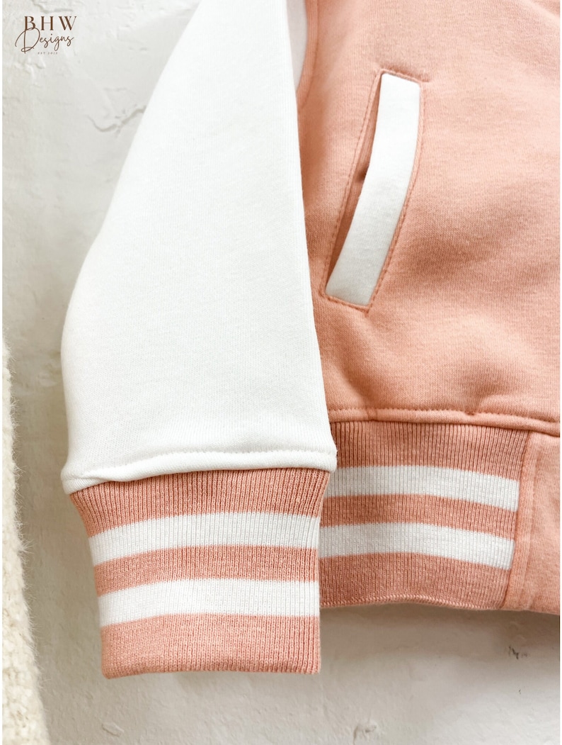 May include: A pink and white baseball jacket with white stripes on the cuffs and waistband. The jacket has a white pocket on the right side.