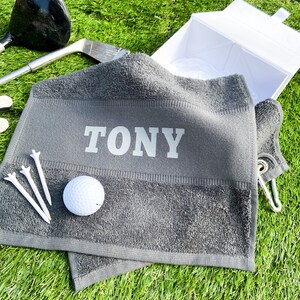 May include: Grey golf towel with the name "TONY" embroidered in white. The towel has a white golf ball and three white golf tees on it.
