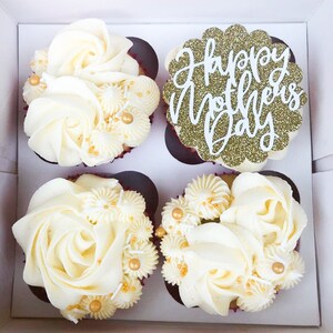Puede incluir: Cuatro cupcakes blancos con glaseado de vainilla decorados con chispas doradas y blancas. El cupcake del centro tiene una decoraci&oacute;n de purpurina dorada que dice "Feliz D&iacute;a de la Madre".