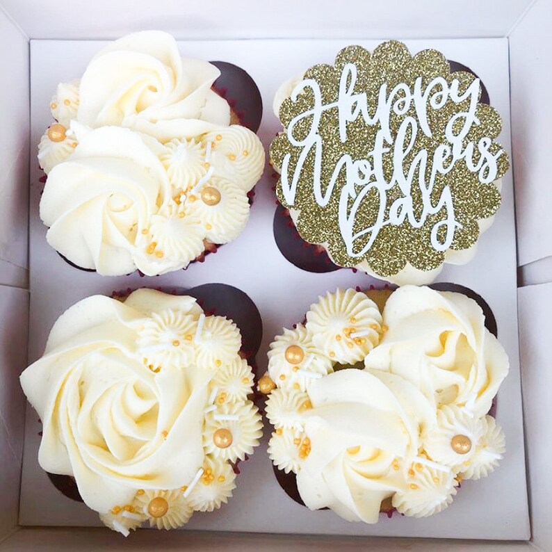 Puede incluir: Cuatro cupcakes blancos con glaseado de vainilla decorados con chispas doradas y blancas. El cupcake del centro tiene una decoraci&oacute;n de purpurina dorada que dice "Feliz D&iacute;a de la Madre".