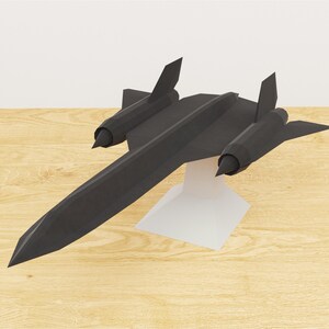 Puede incluir: Un modelo de avión negro SR-71 Blackbird, un avión de reconocimiento, se exhibe sobre un soporte blanco. El modelo tiene un fuselaje largo y delgado, dos motores y estabilizadores verticales distintivos. El avión está sobre un fondo de vetas de madera clara.
