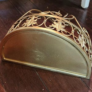 May include: A gold-colored, semi-circular wall-mounted shelf. The shelf has a flat base and a decorative, openwork top with leaf and vine designs. The shelf is made of metal and is likely for decorative purposes.