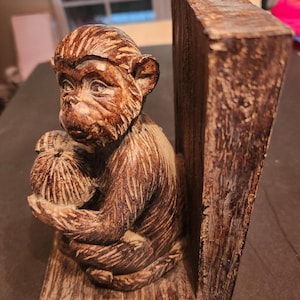 May include: A brown monkey bookend, carved from wood, holding a small sack. The monkey is seated on a wooden base, with a vertical wooden support. The bookend has a rustic, aged appearance, suitable for home decor.