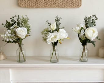 May include: Three glass vases each hold a different arrangement of white flowers and greenery. The vases sit on a white mantelpiece in front of a black fireplace. The background includes macrame wall hangings.