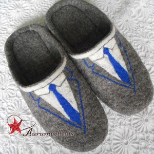 May include: Gray felt slippers with a blue and white embroidered suit design. The slippers have a rounded toe and a closed back.