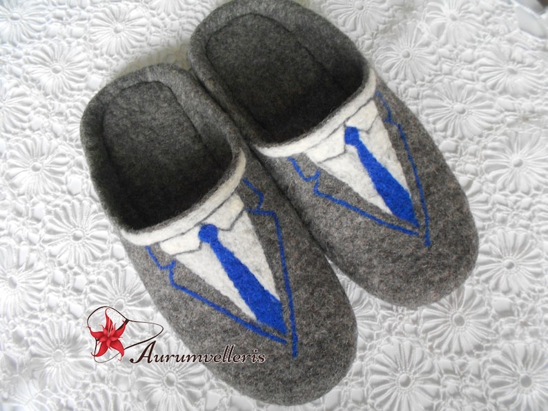 May include: Gray felt slippers with a blue and white embroidered suit design. The slippers have a rounded toe and a closed back.