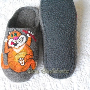 Pu&ograve; includere: Un paio di pantofole in feltro grigio con un design a gatto arancione disegnato. Il gatto tiene un oggetto rosso nella sua zampa e ha un'espressione sorpresa sul suo viso. Le pantofole hanno una suola in gomma nera.