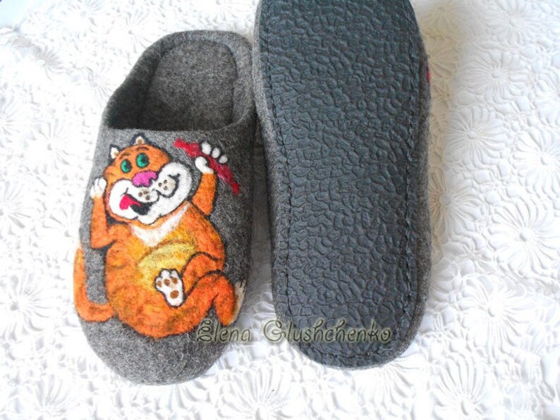 Pu&ograve; includere: Un paio di pantofole in feltro grigio con un design a gatto arancione disegnato. Il gatto tiene un oggetto rosso nella sua zampa e ha un'espressione sorpresa sul suo viso. Le pantofole hanno una suola in gomma nera.