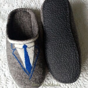 May include: Gray felt slippers with a blue and white embroidered suit and tie design. The slippers have a black rubber sole.