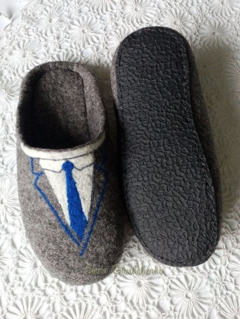 May include: Gray felt slippers with a blue and white embroidered suit and tie design. The slippers have a black rubber sole.