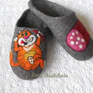 Pu&ograve; includere: Un paio di pantofole in feltro grigio con disegni dipinti a mano. Una pantofola presenta un gatto arancione con una lingua rossa e un fiocco rosso. L'altra pantofola presenta un design a pois rosa e bianco.