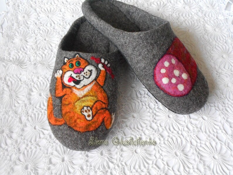 Pu&ograve; includere: Un paio di pantofole in feltro grigio con disegni dipinti a mano. Una pantofola presenta un gatto arancione con una lingua rossa e un fiocco rosso. L'altra pantofola presenta un design a pois rosa e bianco.