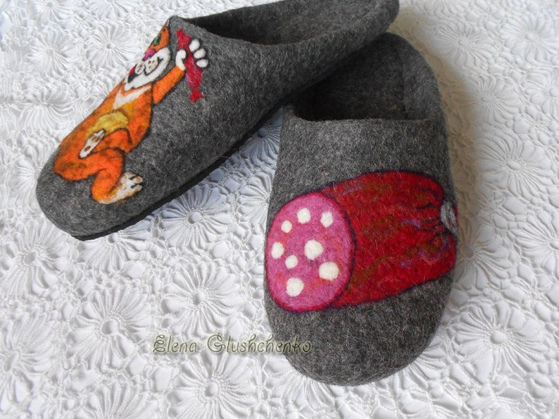 Pu&ograve; includere: Un paio di pantofole in feltro grigio con disegni dipinti a mano. Una pantofola presenta un gatto dei cartoni animati che tiene un salsiccia rossa, mentre l'altra pantofola presenta una salsiccia rossa con pois bianchi.