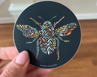 May include: A black and white sticker with a floral bee design. The bee is made up of colorful flowers and leaves. The sticker has the text "HOPE'S PALETTE" at the bottom.