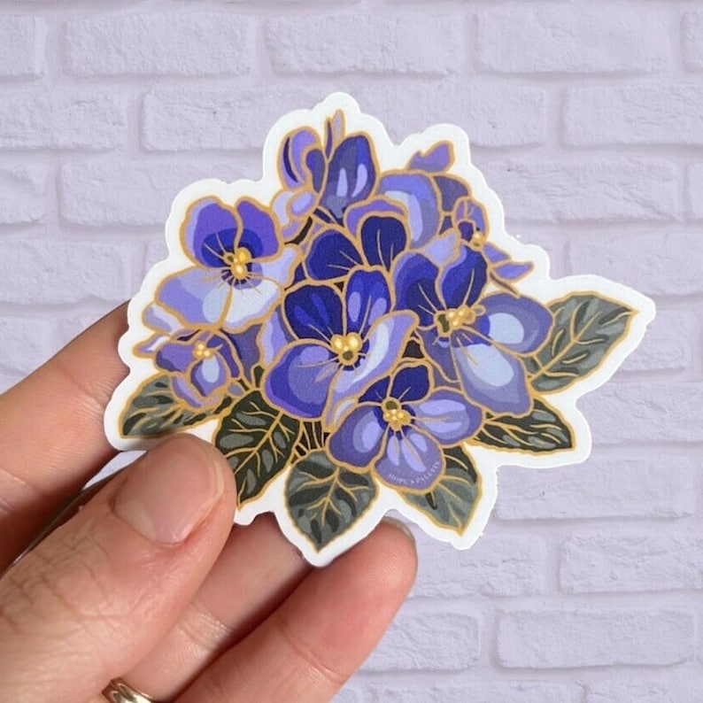 May include: A sticker featuring a cluster of purple and blue flowers with gold accents. The flowers have a watercolor-like appearance and are surrounded by green leaves.